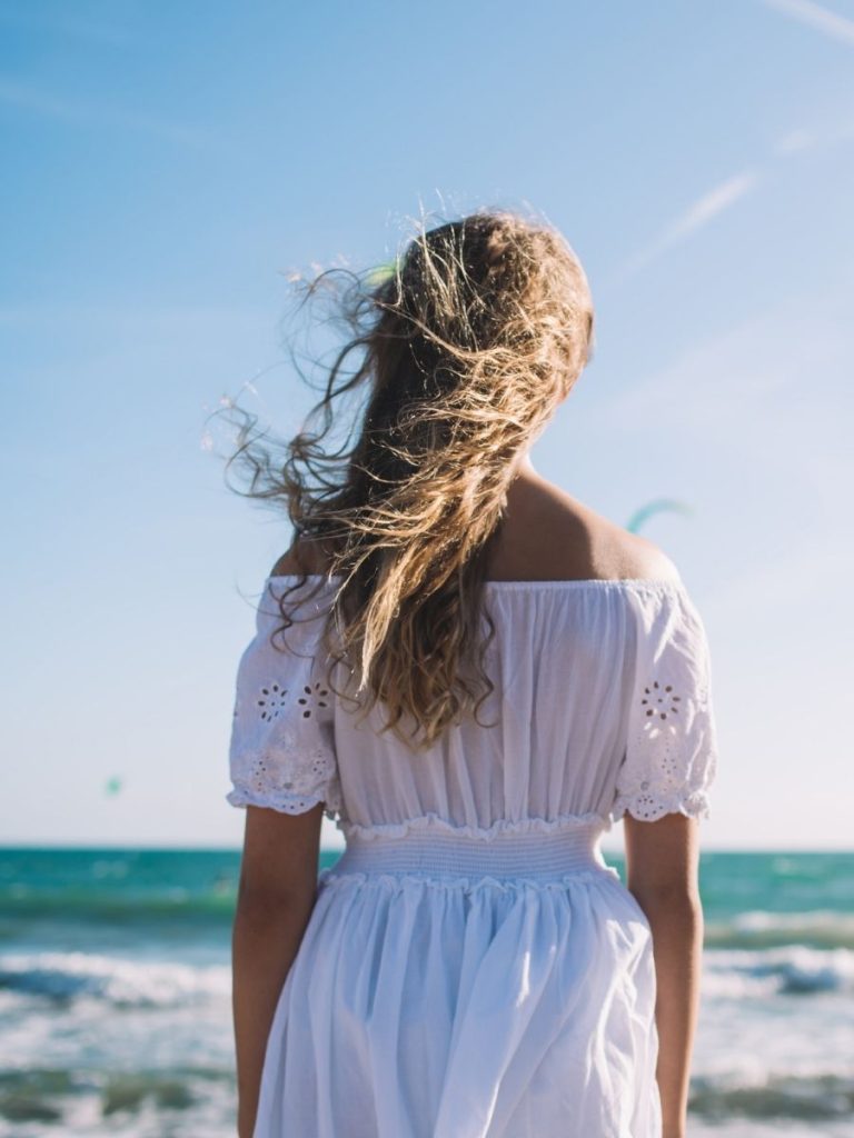 A woman with long, wavy hair stands on the beach facing the ocean, wearing a white off-the-shoulder dress, with wind blowing her hair.
