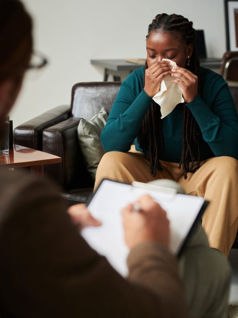 A person sits on a couch, holding a tissue to their face, while another person in the foreground takes notes on a clipboard.