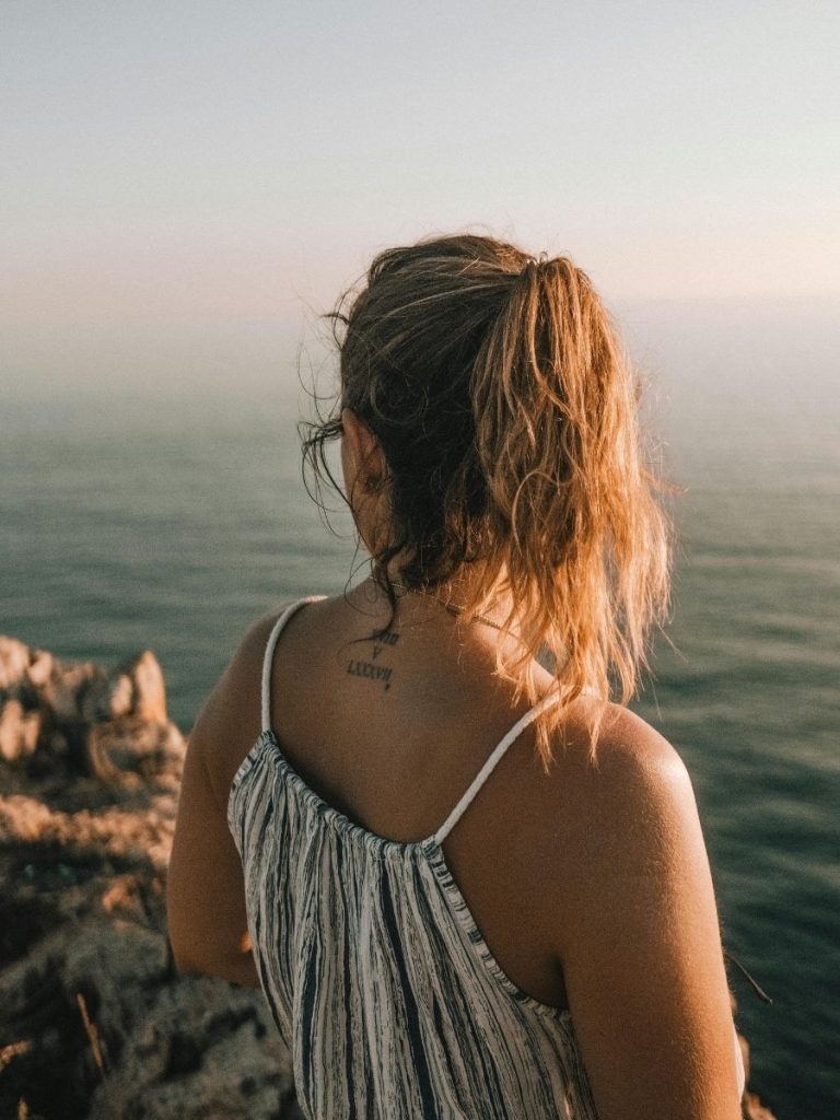 A woman with a ponytail stands on a rocky cliff overlooking the ocean, wearing a striped dress and displaying a tattoo on the back of her neck.